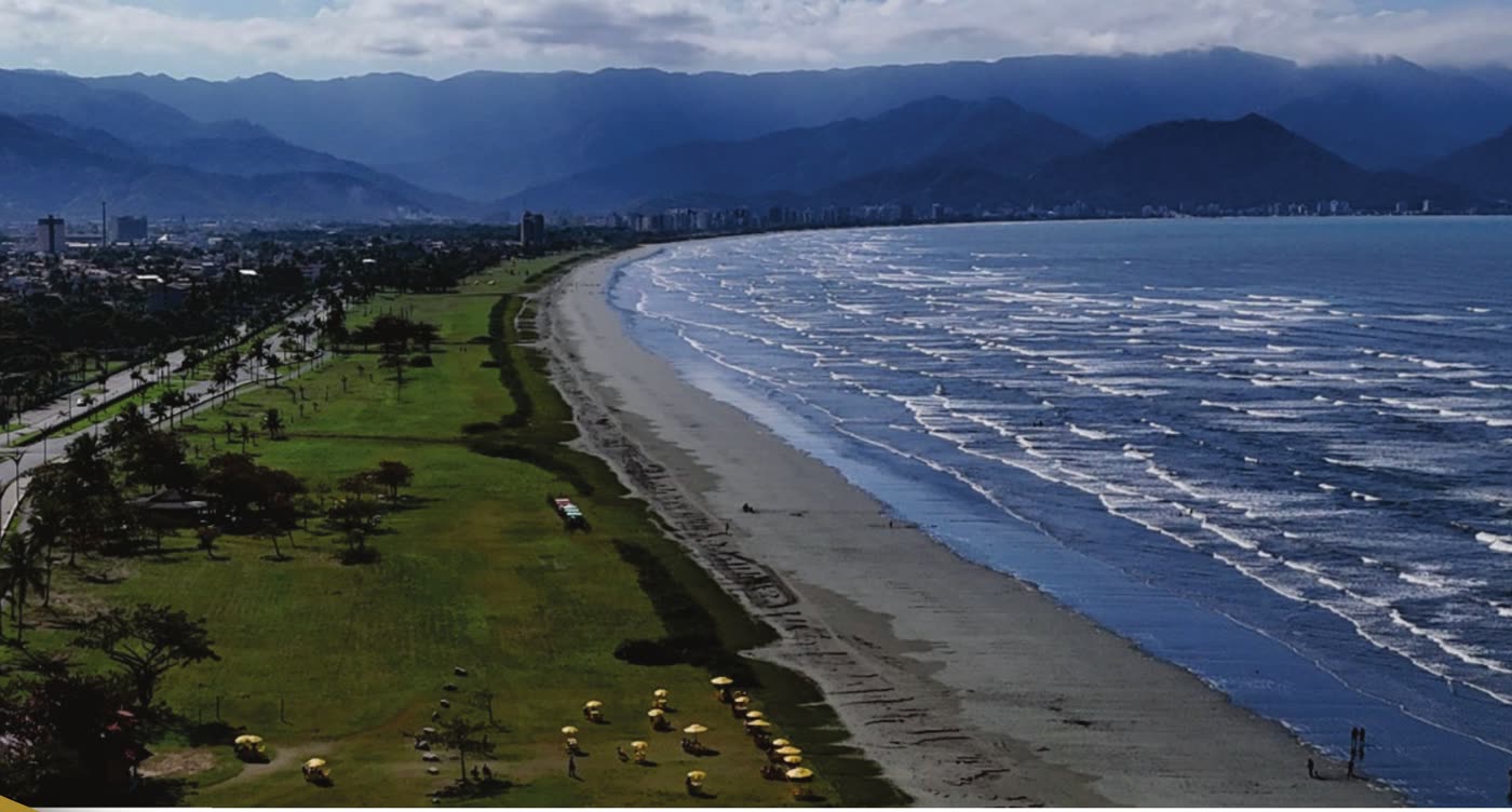 Vista aérea da orla da Praia do Porto Novo, em Caraguatatuba, com o gramado da avenida da praia e a Serra do Mar ao fundo